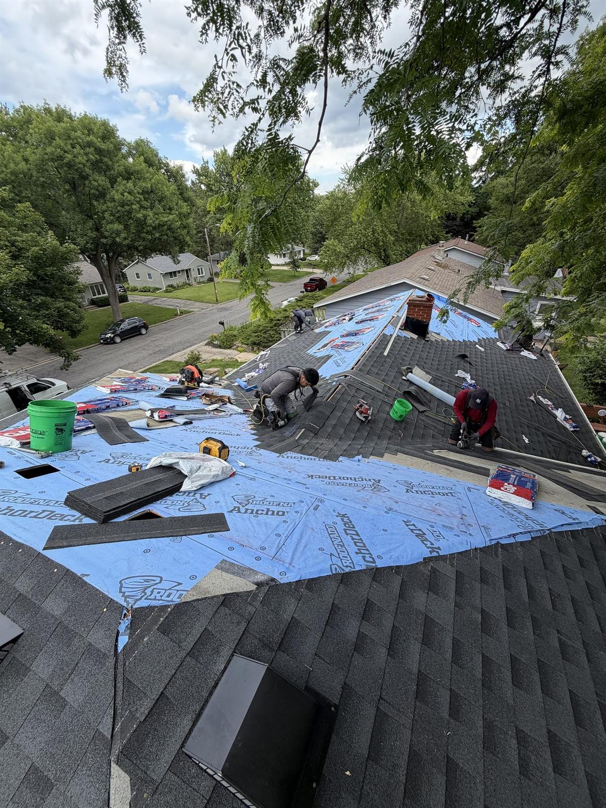 Roofing crew installing architectural shingles over blue synthetic underlayment on a steep residential roof