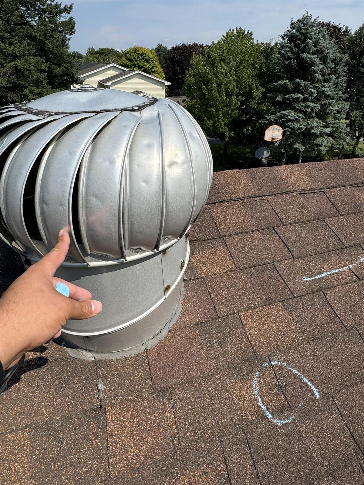 Hail-dented turbine attic vent held during a roof inspection with chalk-marked hail impacts on the shingles behind it